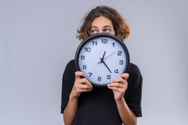 Kerl mit langen Haaren im schwarzen T-Shirt, das eine Wanduhr auf weißer Wand hält