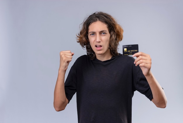 Kerl mit langen Haaren im schwarzen T-Shirt, der eine Bankkarte auf weißer Wand hält