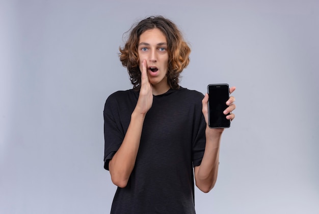 Kerl mit langen Haaren im schwarzen T-Shirt hält ein Telefon und flüstert auf weißer Wand