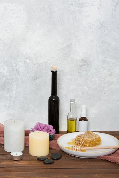 Kerzen Letzter; Rose; wesentliche Flasche; Bienenwabe und Schöpflöffel auf hölzernem Schreibtisch gegen graue Wand