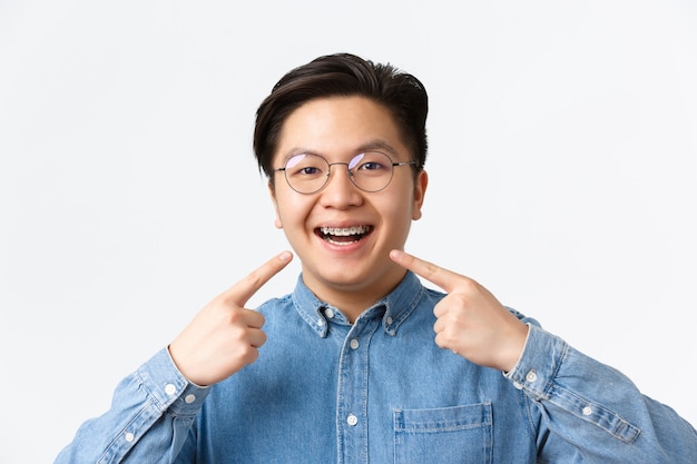 Kieferorthopädie und Stomatologie-Konzept. Nahaufnahme eines zufriedenen asiatischen Kerls, Zahnklinik-Kunde, der glücklich lächelt und auf seine Zahnspangen zeigt, weißer Hintergrund steht, Qualität empfehlen.