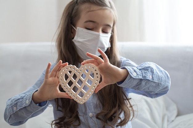Kind, das medizinische schützende Gesichtsmaske zum Gesundheitsschutz von Coronavirus trägt, hält hölzernes Herz.