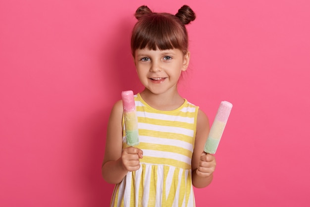 Kind, das zwei eiscreme hält, glückliches kleines mädchen, das wassereis genießt, niedliches kind, das köstliches straßenessen im sommer schmeckt, lokalisiert über rosa wand.