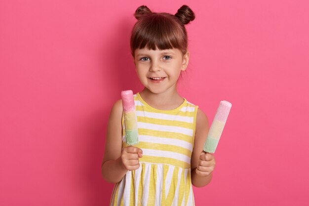 Kind, das zwei Eiscreme hält, glückliches kleines Mädchen, das Wassereis genießt, niedliches Kind, das köstliches Straßenessen im Sommer schmeckt, lokalisiert über rosa Wand.
