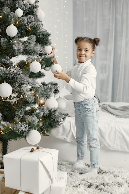 Kind in einem weißen Pullover. Tochter, die nahe Weihnachtsbaum steht.