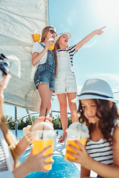 Kinder an Bord der Yacht trinken Orangensaft. Teenager- oder Kindermädchen gegen blauen Himmel im Freien. Farbenfrohe Kleider.