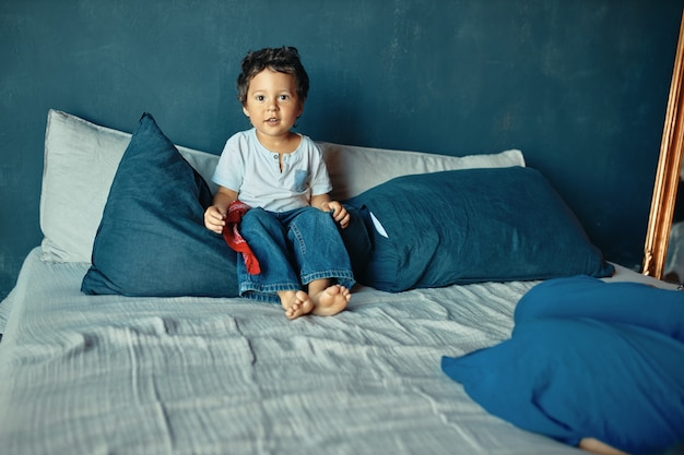 Kinder-, Bett- und Erziehungskonzept. Netter kleiner Junge der gemischten Barfußrasse, der auf Bett sitzt und bereit ist, nach dem Tagesschlaf zu spielen.