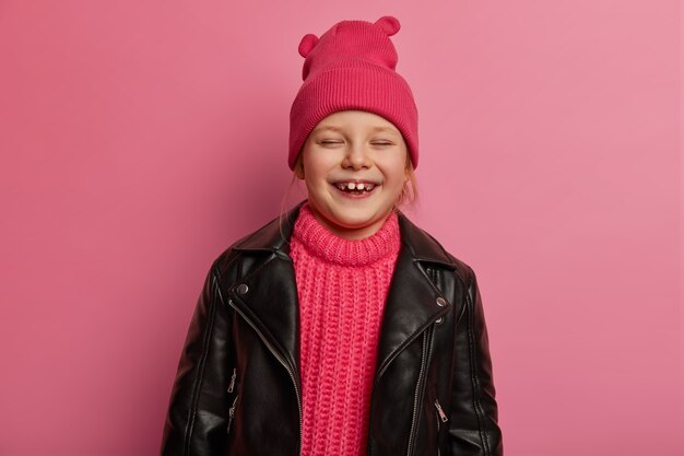 Kinder, glückliche Gefühle und aufrichtige Gefühle Konzept. Überglückliches kleines süßes Mädchen lacht, spielt mit den Eltern herum, trägt Hut, Strickpullover und Schaumjacke, drückt Freude und Glück aus