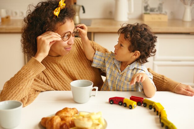 Kinder, Kinder, glückliche Kindheit, Familienbande und Elternkonzept. Bild der attraktiven jungen hispanischen Frau, die Kaffee am ktichen Tisch trinkt und lächelt, während Säuglingssohn ihre Brille abnimmt