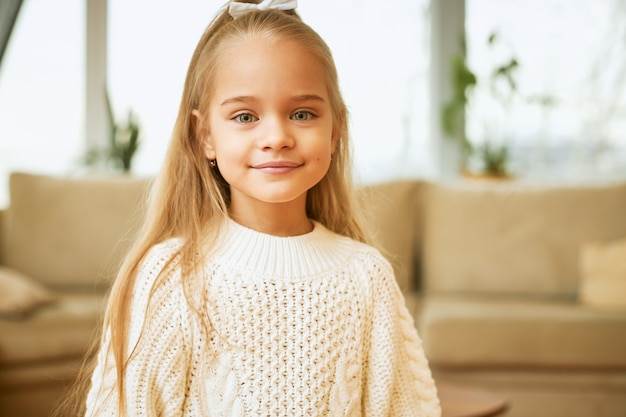 Kinder, Schönheit und Stil. Schönes kaukasisches kleines Mädchen mit den blauen Augen, dem niedlichen Lächeln und den langen Haaren, die im Wohnzimmer posieren, gekleidet im kuscheligen weißen Pullover, in der guten Laune, im freudigen Blick