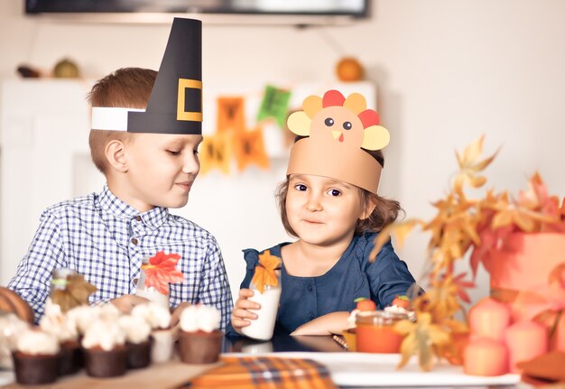 Kinder sitzen auf festlichem tisch und feiern thanksgiving. kinder in papiertruthahnhut und pilgerhüten, die cupcakes essen und milch trinken.