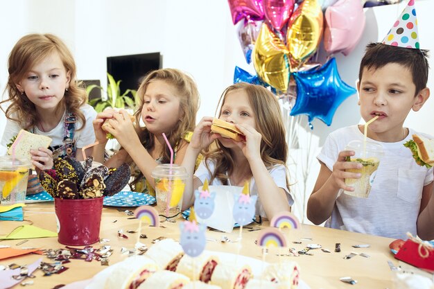 Kinder- und Geburtstagsdekorationen. Jungen und Mädchen am Tisch mit Essen, Kuchen, Getränken und Party-Gadgets.