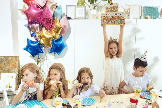 Kinder- und Geburtstagsdekorationen. Jungen und Mädchen am Tisch mit Essen, Kuchen, Getränken und Party-Gadgets.