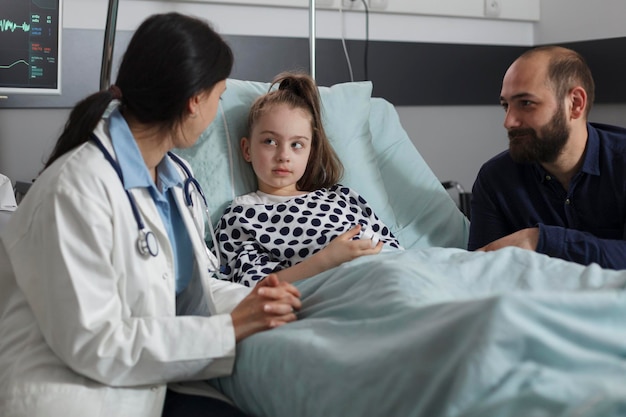 Kinderarzt diskutiert mit kranker kleiner Tochter, die im Patientenbett ruht, während der fürsorgliche Vater neben ihr sitzt. Medizinischer Experte der pädiatrischen Gesundheitseinrichtung, der den Gesundheitszustand von unwohlem Mädchen berät.