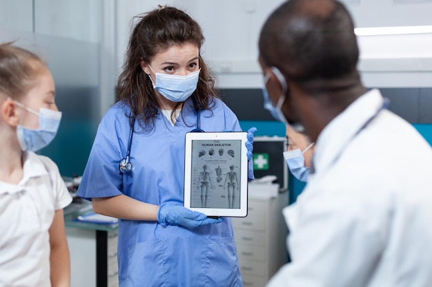 Kinderarztkrankenschwester mit schützender Gesichtsmaske gegen covid19, die einen Tablet-Computer mit Knochenradiographie auf dem Bildschirm hält. Afroamerikanischer Arzt, der medizinisches Fachwissen im Krankenhausbüro erklärt