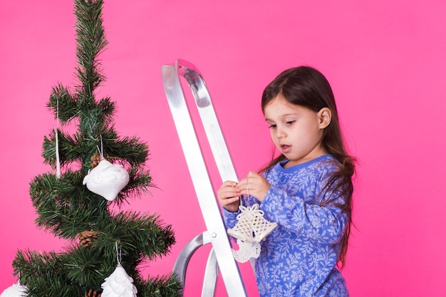 Kinderferien und weihnachtskonzept kleines mädchen, das weihnachtsbaum auf rosa hintergrund verziert