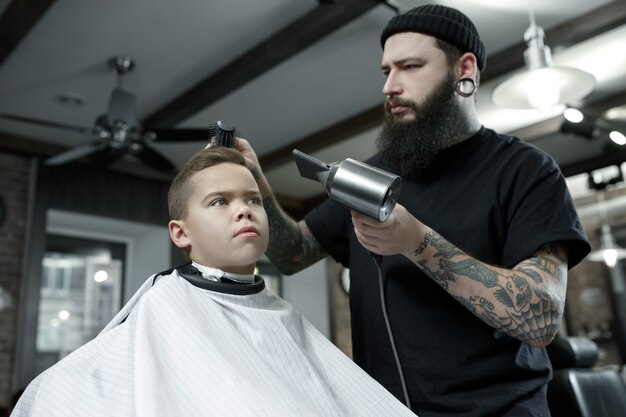 Kinderfriseur, der kleinen Jungen gegen einen dunklen Hintergrund schneidet.