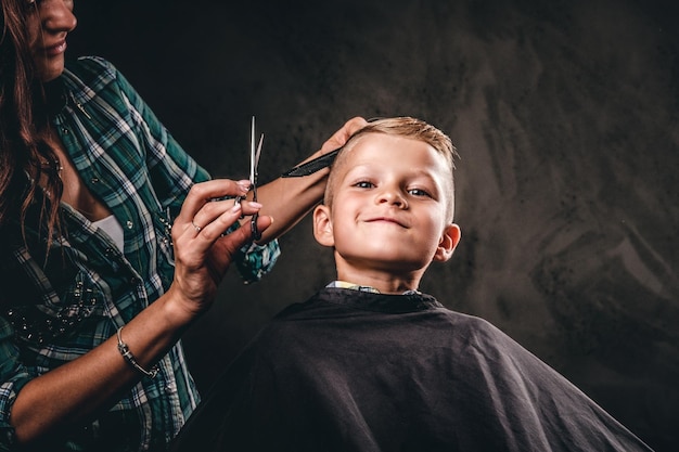 Kinderfriseur mit schere schneidet kleinen jungen vor dunklem hintergrund. zufriedener süßer vorschuljunge, der einen haarschnitt bekommt.
