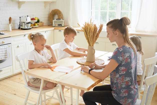 Kindheit, Familie, Kreativität, Freizeit und Hobbykonzept. Horizontale Aufnahme einer jungen, lässig gekleideten Mutter, die mit ihren drei Kindern Mutterschaftsurlaub verbringt und gemeinsam Origami-Kunsthandwerk macht