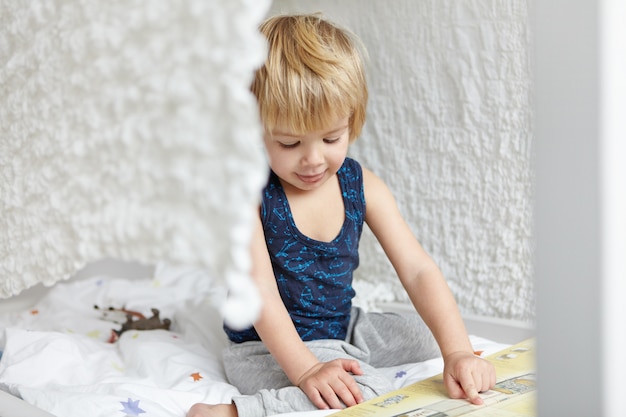 Kindheit und Freizeit. Entzückender süßer kleiner blonder Junge im Schlafanzug, der auf seinem Bett vor offenem Buch sitzt, seinen Zeigefinger zeigt, Bilder zeigt, konzentriert aussieht.