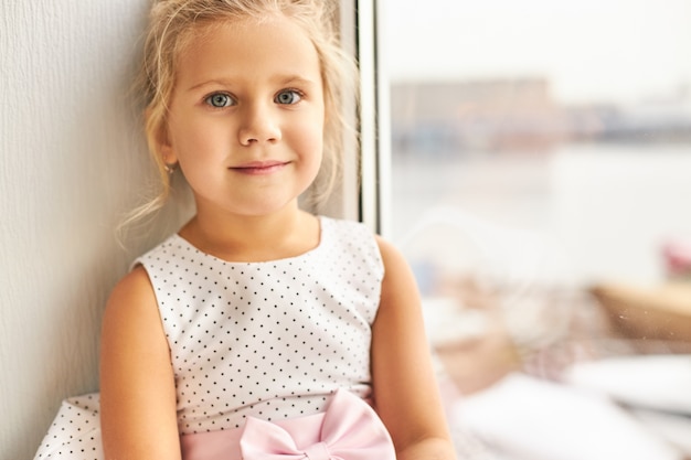 Kindheit und unschuldiges Konzept. Porträt des charmanten niedlichen kleinen Mädchens mit gesammeltem hellem Haar und großen schönen Augen, die am Fenster sitzen, glücklichen Gesichtsausdruck und lächelnd haben