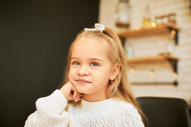 Kindheitskonzept. Innenbild des schönen kleinen Mädchens mit dem langen Haar, das in der Küche mit Girlande sitzt, die weißes Band und gestrickten Pullover trägt, Hand unter ihrem Kinn hält und lächelt