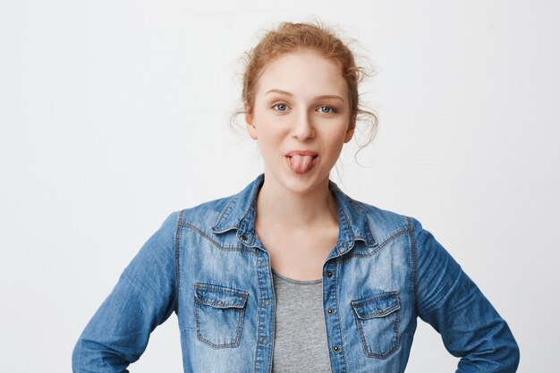 Kindische emotionale rothaarige Teenager-Mädchen, die Zunge herausstrecken und lächeln, machen Gesichter, während sie mit den Händen auf der Taille stehen