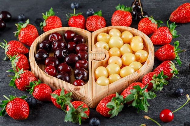 Kirschen mit Erdbeeren, Blaubeeren in einer Holzplatte auf grauer Oberfläche, hohe Winkelansicht.