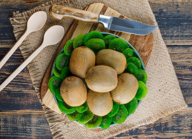 Kiwi mit getrockneten Scheiben, Schneidebrett, Messer, Löffeln in einem Teller auf Holz und Stück Sackhintergrund, flach liegen.