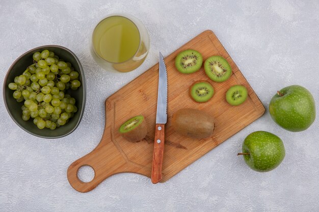 Kiwi-Scheiben der Draufsicht mit einem Messer auf einem Schneidebrett mit grünen Äpfeln und Trauben in einer Schüssel mit Apfelsaft in einem Glas auf einem weißen Hintergrund