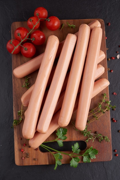 Klassische Schweinefleischwürste mit gekochtem Fleisch auf Schneidebrett mit Pfeffer und Basilikum, Petersilie, Thymian und Kirschtomaten.