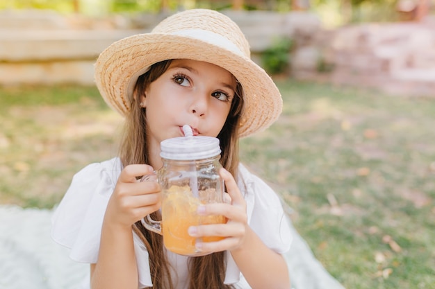 Kleine Dame mit braunen Augen und langen schwarzen Wimpern, die weg schauen, während Fruchtsaft trinken. Nettes Mädchen, das Glas des Cocktails hält und dieses kalte Getränk im Park während des Urlaubs genießt.