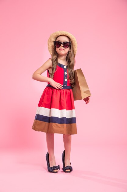 kleine Fashionista mit einer Einkaufstasche in einem Sommerhut und einer Brille, auf einem farbigen rosa Hintergrund in Mamas Schuhen, das Konzept der Kindermode