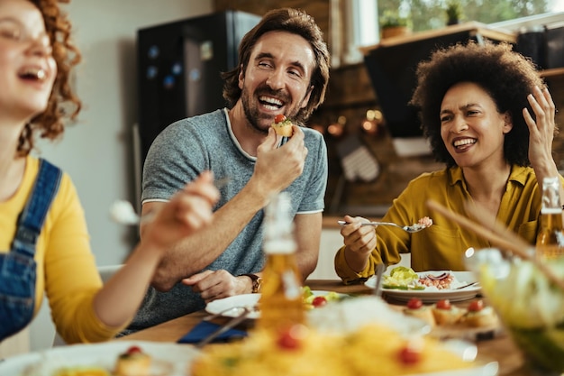 Kleine Gruppe junger glücklicher Menschen, die das Mittagessen genießen, während sie sich am Esstisch unterhalten