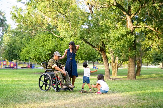 Kleine Kinder arrangieren Brennholz für Lagerfeuer im Freien in der Nähe von Mutter und behinderten Militärvater im Rollstuhl. Behinderte Veteran oder Familie im Freien Konzept