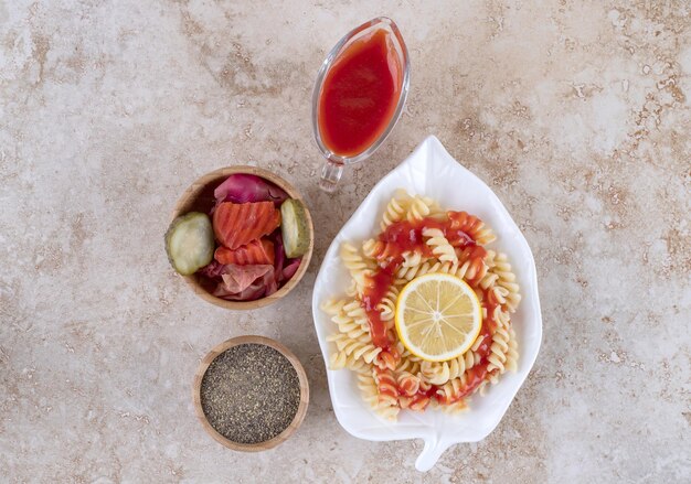 Kleine Pastaplatte, Zitronenscheibe, verschiedene Essiggurken und eine Schüssel mit schwarzem Pfeffer auf Marmoroberfläche.