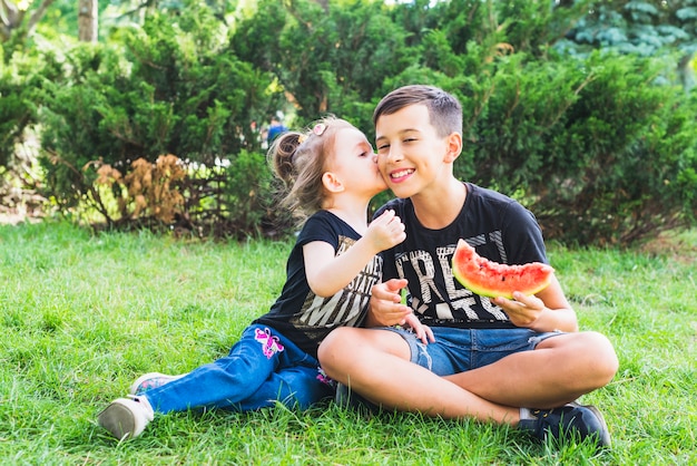 Kleine Schwester, die ihren Bruder hält Wassermelonenscheibe küsst