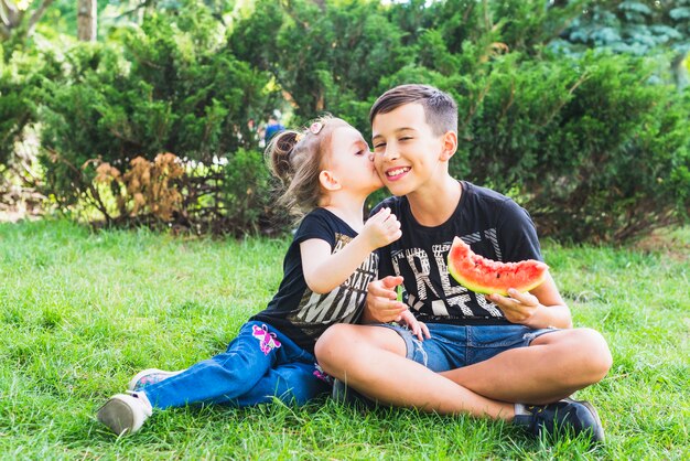 Kleine Schwester, die ihren Bruder hält Wassermelonenscheibe küsst