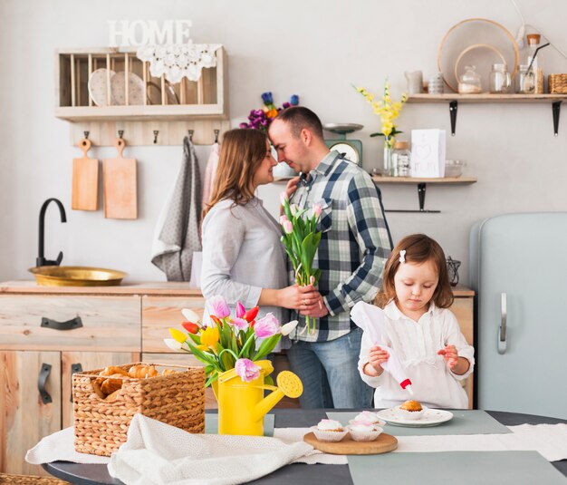 Kleine Tochter, die kleinen Kuchen nahe Eltern mit Blumen macht