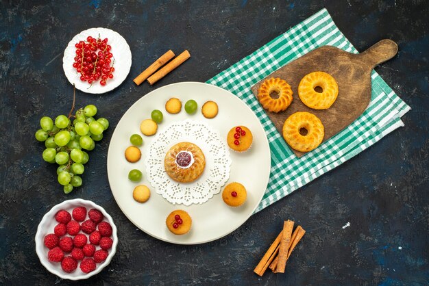 Kleine Torte der oberen entfernten Ansicht mit Sahnekeksen und Himbeeren und Preiselbeeren der grünen Trauben auf dem dunklen Schreibtisch süß