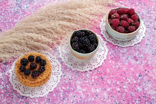 Kleiner Brombeerkuchen mit halber Draufsicht mit Himbeeren und frischen Brombeeren auf dem hellrosa Hintergrund.