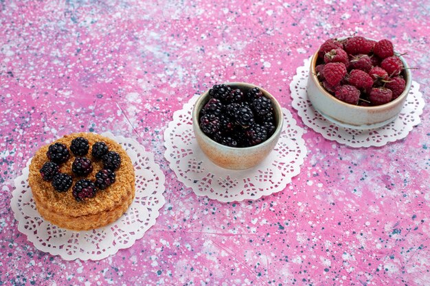 Kleiner Brombeerkuchen mit halber Draufsicht mit Himbeeren und frischen Brombeeren auf dem hellrosa Hintergrund.