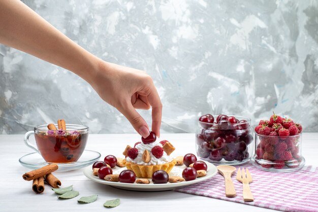 kleiner cremiger Kuchen mit Himbeerkirschen und kleinen Keksen Tee Zimt auf Weißlicht Schreibtisch, Frucht süße Beerencreme