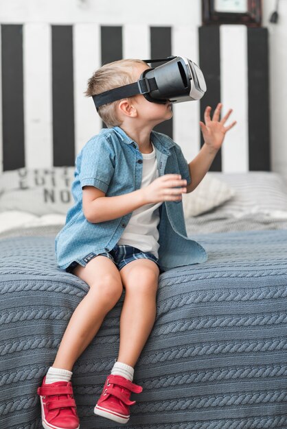 Kleiner Junge, der auf Bett unter Verwendung der Schutzbrillen der virtuellen Realität sitzt