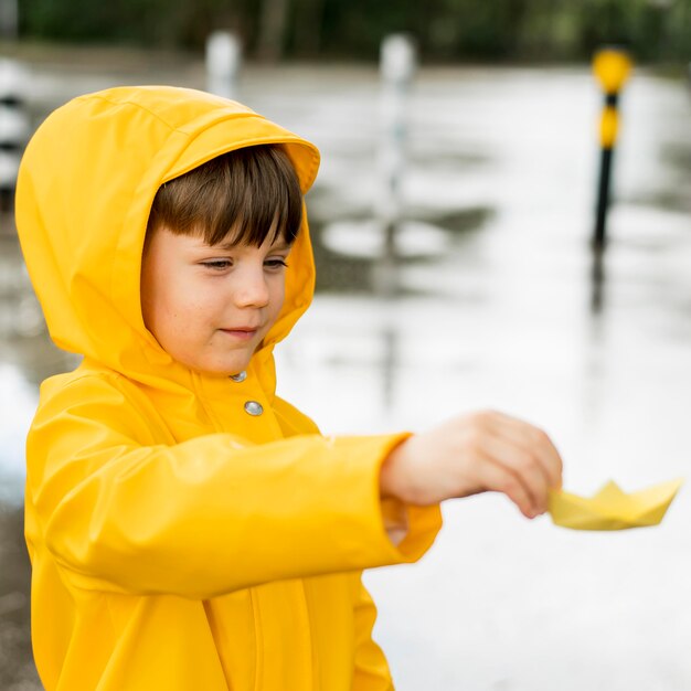Kleiner Junge, der im Regen mit einem Papierboot spielt