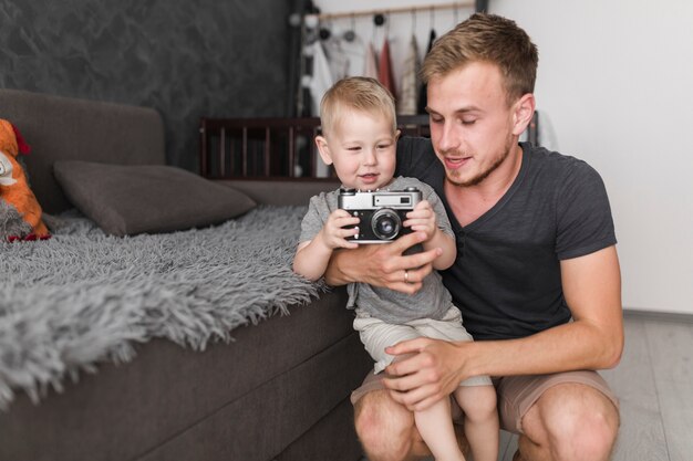 Kleiner Junge, der in der Hand auf dem Schoss seines Vaters hält Kamera sitzt