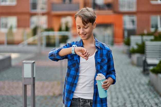Kleiner Junge, der mit blauer Smartwatch spricht und Tee in der Nähe der Schule trinkt.