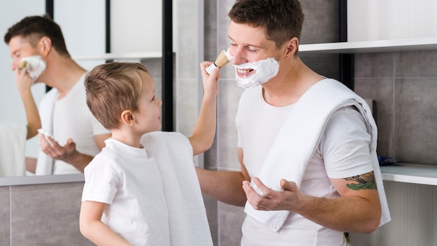 Kleiner Junge, der Schaumgummi auf Gesicht seines Vaters mit Bürste im Badezimmer rasiert