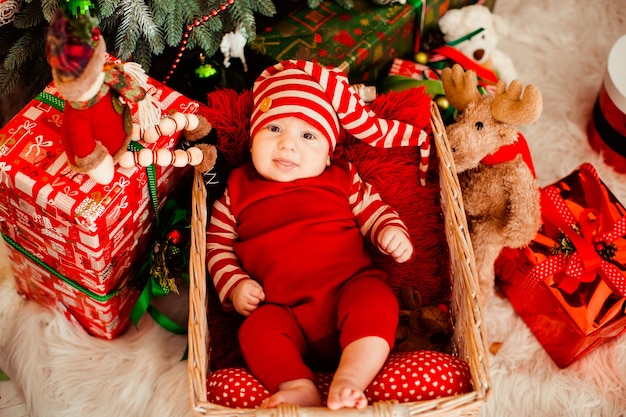 Kleiner Junge in der lustigen roten Klage und im langen Hut liegt im Korb vor einem Weihnachtsbaum