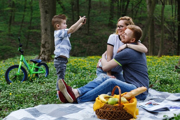 Kleiner Junge macht ein Foto seiner Eltern auf dem Smartphone während eines Picknicks im Park